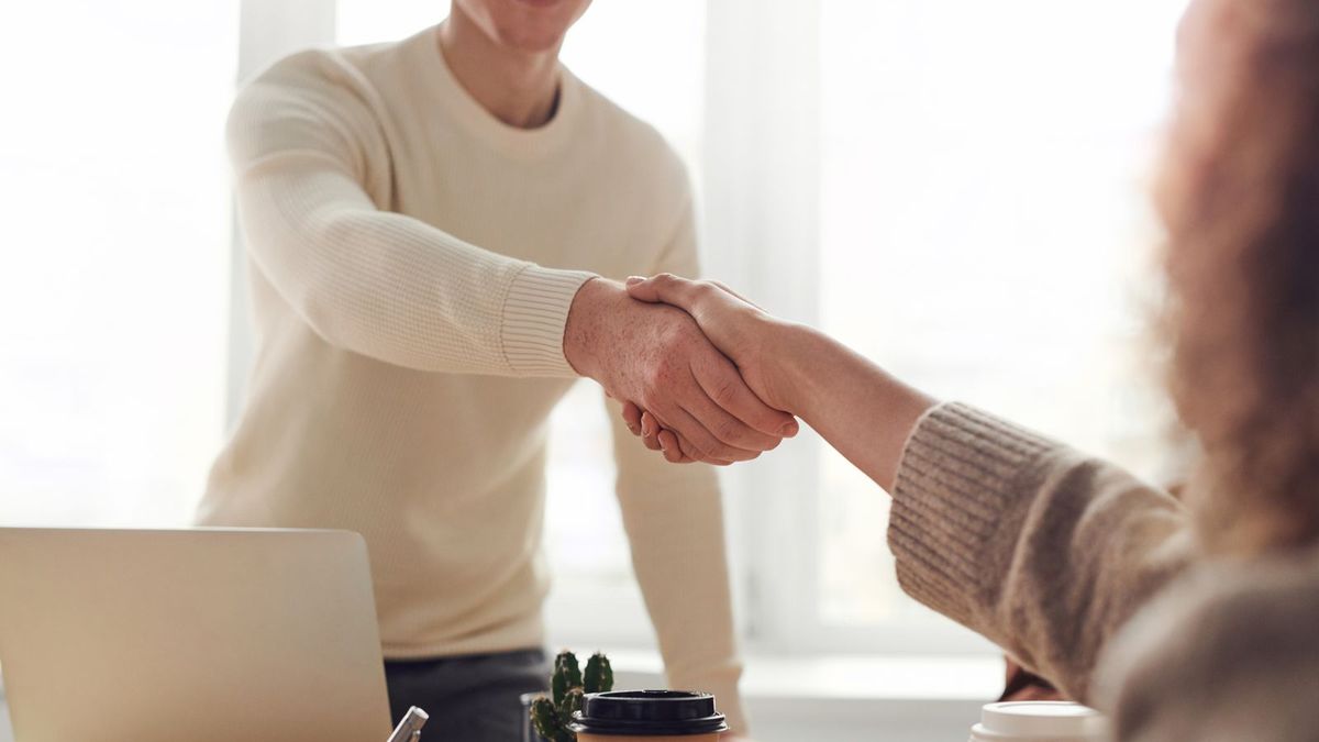 Zwei Personen geben sich am Besprechungstisch die Hand, im Vordergrund Unterlagen und Kaffeetassen