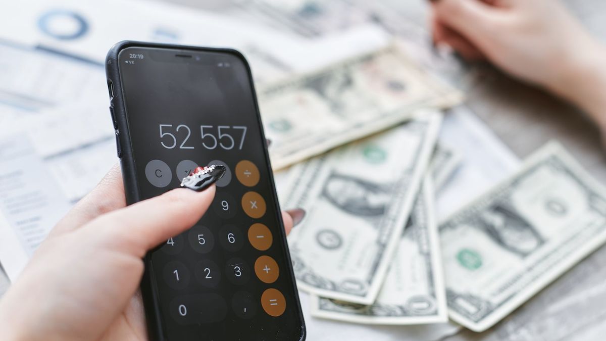 Hände tippen Beträge in den Rechner eines Smartphones, daneben liegen Geldscheine auf dem Tisch